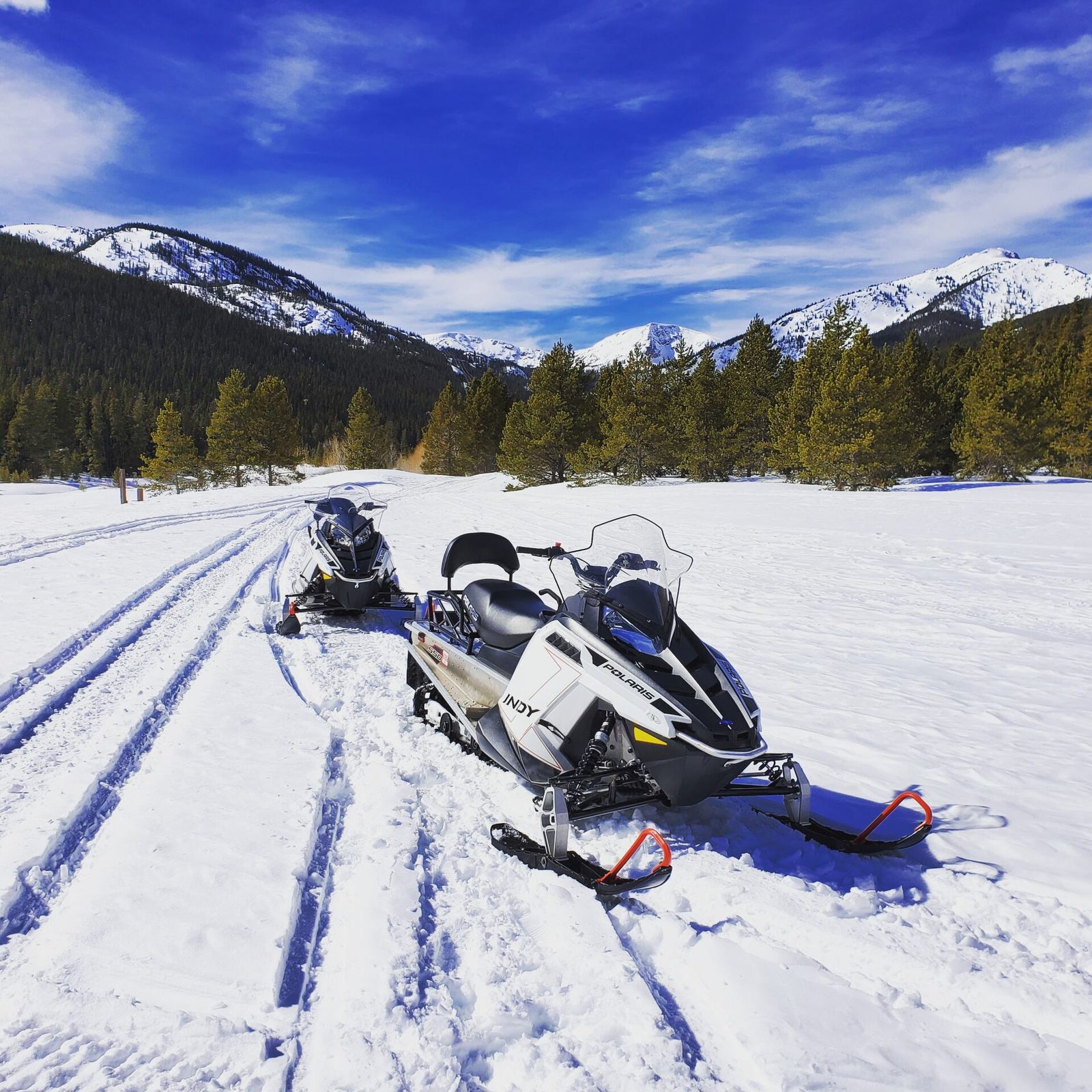 Snowmobiles on a groomed trail under a deep blue sky with snowy peaks behind